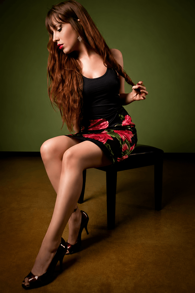 A woman with long brown hair sits sideways on a chair, wearing a black tank top and a floral skirt. She holds a strand of her hair while posing elegantly against a green backdrop.