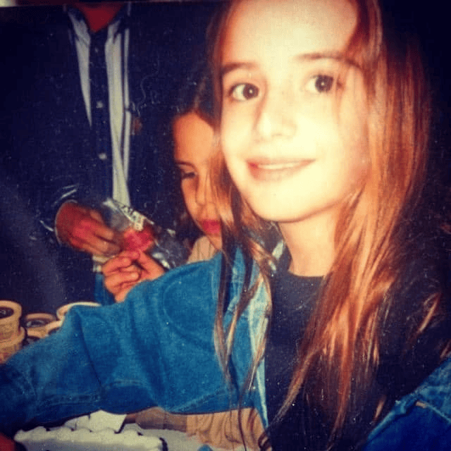 A young girl with long brown hair and a smile, sitting at a table with someone in the background. She is wearing a blue denim jacket.